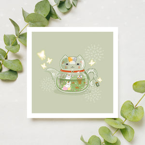 A square print with an imaginative illustrated clear teapot in the shape of a cat. Inside the clear teapot there are cats playing in the green tea. The teapot is set against a soft, earthy green with butterfly and fractal designs. The print is displayed on a table with eucalyptus leaves.